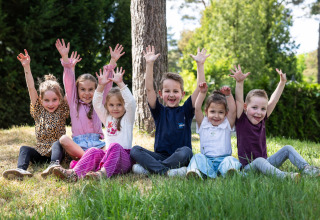 A group of happy children sit on the grass with raised hands at Campsite Roland in Limburg, Netherlands.