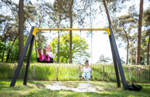 Two children are playing on swings at a playground surrounded by trees in Campsite Roland, Limburg, Netherlands.