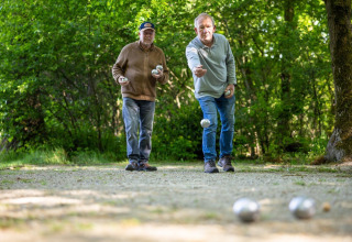 Two older men play pétanque outdoors on a trail surrounded by trees at Campsite Roland in Limburg, Netherlands.