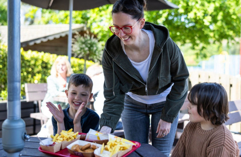 A woman serves trays of fries and nuggets to two happy children at an outdoor campsite dining area.