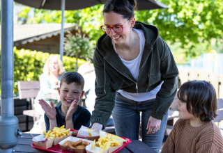 A woman serves trays of fries and nuggets to two happy children at an outdoor campsite dining area.