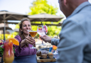 Two people toast with colorful cocktails on the outdoor terrace at Campsite Roland in Limburg, Netherlands.