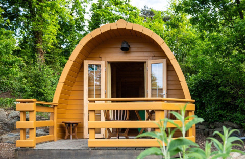 A small wooden pod tiny house with a veranda, surrounded by lush green trees and nature.