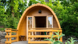 A small wooden pod tiny house with a veranda, surrounded by lush green trees and nature.
