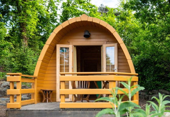 A small wooden pod tiny house with a veranda, surrounded by lush green trees and nature.
