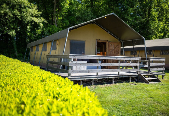 Safari-Zelt mit überdachter Holzterrasse im Grünen, umgeben von Bäumen und Hecken, bei Tageslicht.
