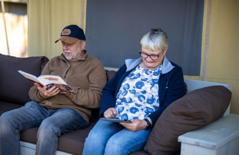 To ældre voksne sidder afslappet i en safaritelt på Campingplads Roland, Holland, og nyder bøger og tablet.