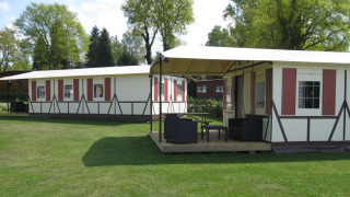 Two half-timbered safari tents on a green lawn, surrounded by trees under a sunny sky with scattered clouds.