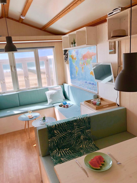 Lyst strandhytteinteriør med verdenskort, pastelfarvede sofaer og udsigt til stranden gennem vinduet.