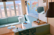 Interior luminoso de cabaña playera con sofás pastel, mapa mundial y vista al mar a través de la ventana.