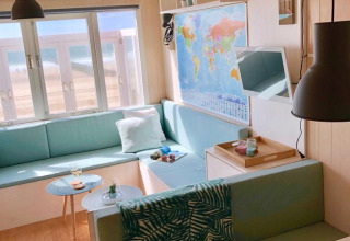 Bright beach cabin interior with pastel seating, world map on the wall, and seaside view from the window.