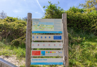 Cartel informativo en la playa de Dishoek, Países Bajos, con mapa, reglas y consejos rodeado de vegetación.
