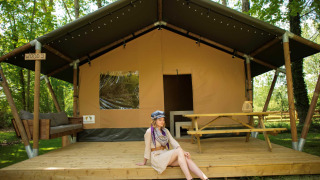 Woman sits on the deck of Safari tent WOOD, with picnic table and sofa in a wooded campsite.