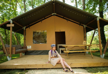 Woman sits on the deck of Safari tent WOOD, with picnic table and sofa in a wooded campsite.