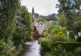 Historisches Gebäude mit Turm am Fluss, umgeben von üppigem Grün im Le Hameau de l'Ourthe, Belgien.