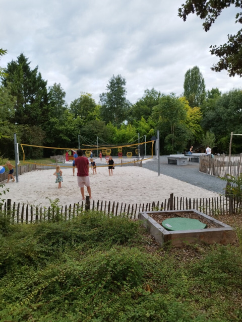 Bambini e adulti giocano a pallavolo su un campo di sabbia in un campeggio circondato da alberi verdi.
