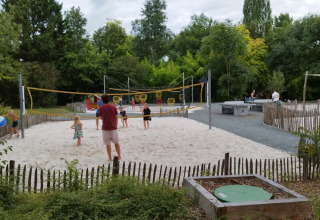 Bambini e adulti giocano a pallavolo su un campo di sabbia in un campeggio circondato da alberi verdi.