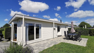 Photo du Chalet Basic, un chalet blanc avec terrasse, mobilier de jardin et ciel bleu avec nuages.