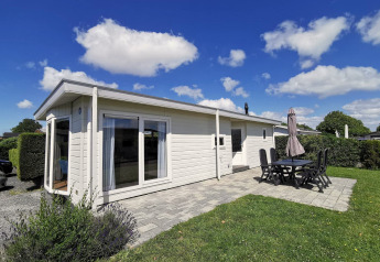 Foto af Chalet Basic, en hvid feriebolig med terrasse, havemøbler og blå himmel med skyer.