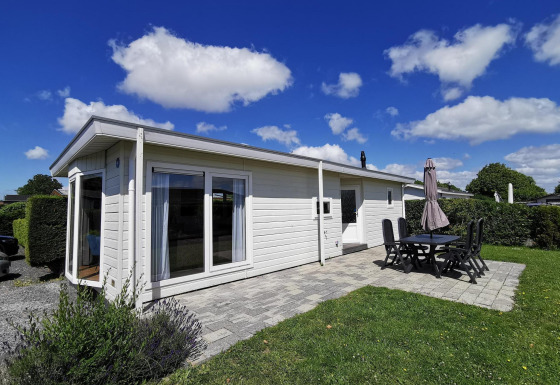 Photo du Chalet Basic, un chalet blanc avec terrasse, mobilier de jardin et ciel bleu avec nuages.