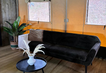 Safari-Zelt-Innenraum mit schwarzem Sofa, kleinem Tisch, Vase, Pflanze und Holzfußboden, Fenster hinten.