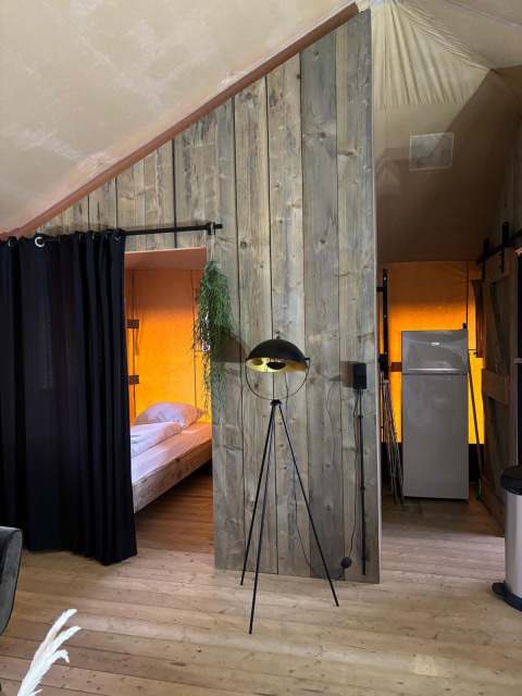 Binnenzicht van een safaritent met houten vloer, bed achter een zwart gordijn en koelkast achteraan.
