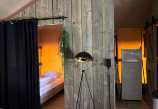 Binnenzicht van een safaritent met houten vloer, bed achter een zwart gordijn en koelkast achteraan.
