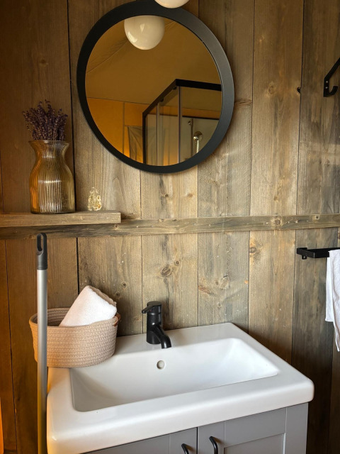 Badkamer in safaritent met houten wand, ronde spiegel, modern wastafel, handdoeken en decoratievaas.