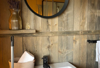 Badkamer van safaritent met houten muur, ronde spiegel, witte wastafel en een decoratieve vaas.