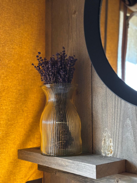 Een glazen vaas met gedroogde lavendel op een plank in een safaritent, gehuld in warm geel licht.