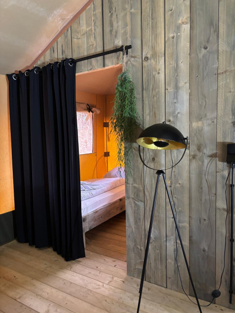 Binnenzicht van een safaritent met houten wanden, zwart gordijn, bed en moderne vloerlamp.