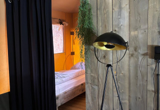 Interieur van een safaritent met bed, zwart gordijn, houten wanden en een moderne vloerlamp.