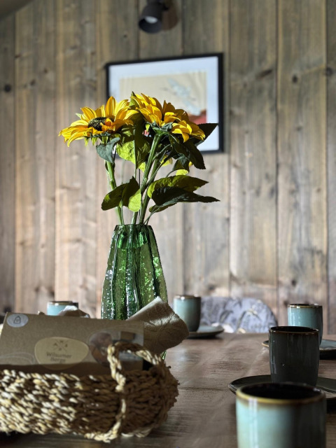 Gele zonnebloemen in groene vaas op houten tafel met bekers, mand en houtwand in safaritent.
