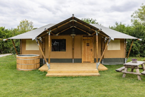 Safari tent Glamfield Lodge II met houten hottub, veranda en picknickbank op een groene grasweide.