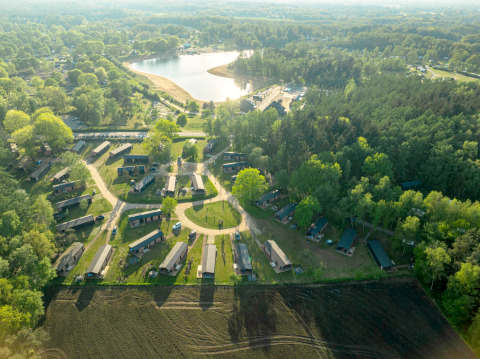Luchtfoto van een glampingverblijf met hutten, gelegen bij een meer en omgeven door groen bos.