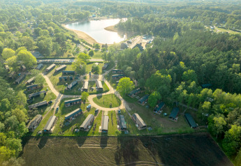 Aerial view of a glamping site with cabins, surrounded by forest, fields, and a scenic lake nearby.