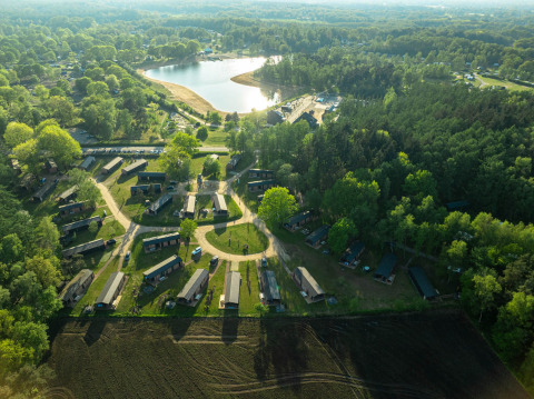 Aerial view of a glamping site with cabins, surrounded by forest, fields, and a scenic lake nearby.