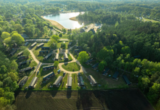 Aerial view of a glamping site with cabins, surrounded by forest, fields, and a scenic lake nearby.