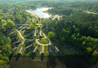 Luftfoto af glampingområde med hytter og natur tæt på sø og skov, omgivet af grønne træer.