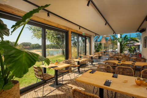 Licht glampingrestaurant met rotan stoelen, grote planten en panoramisch uitzicht over het meer.