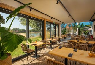 Licht glampingrestaurant met rotan stoelen, grote planten en panoramisch uitzicht over het meer.