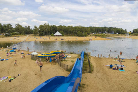 Zandstrand met blauwe glijbaan bij meer, mensen en glamping voorzieningen in een bosrijke omgeving.