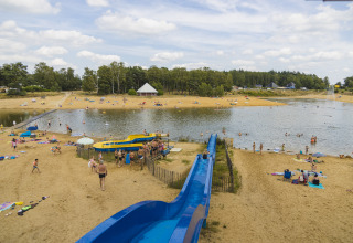 Zandstrand met blauwe glijbaan bij meer, mensen en glamping voorzieningen in een bosrijke omgeving.