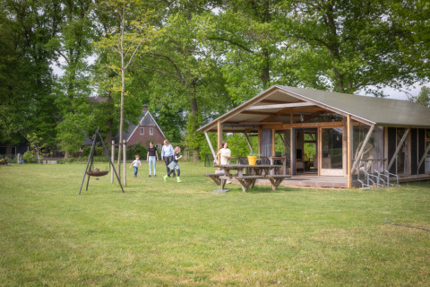 Gezin geniet buiten bij een Luxe lodge met veranda en ruime groene tuin omringd door bomen.