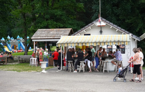 Mensen staan en zitten aan een kraam op Family's Camping Le Savoy, Auvergne-Rhône-Alpes, met speeltuin nabij.