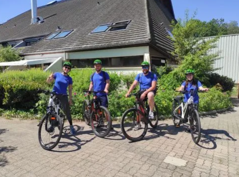 Four people in blue shirts and green helmets on bicycles in front of Family's Camping Le Savoy, France.