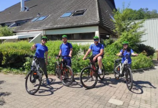 Cuatro personas con camisetas azules y cascos verdes en bicicleta frente a Family's Camping Le Savoy, Francia.