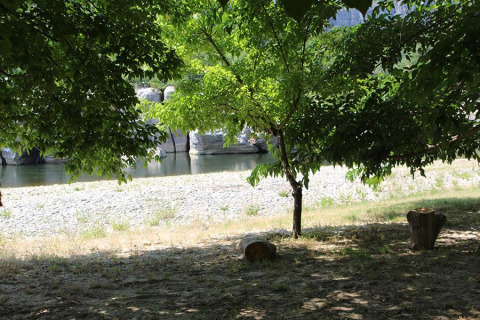 Zona sombreada bajo árboles con vista al río y playa de piedras en Camping Paradis Family des Issoux, Francia.