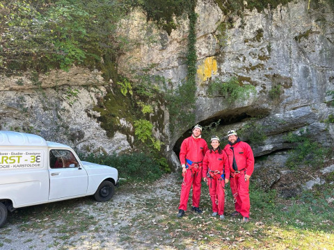 Tre personer i røde dragter foran en grotte med en hvid varevogn ved Family's Camping Le Savoy, Frankrig.