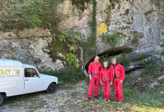 Tre personer i røde dragter foran en grotte med en hvid varevogn ved Family's Camping Le Savoy, Frankrig.